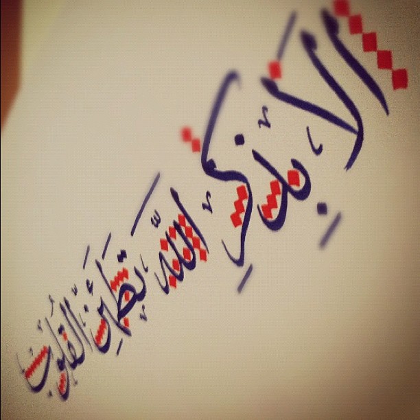 La aleya «ألا بذكر الله تطمئن القلوب» caligrafiada en tinta azul oscuro con puntos rojos, en diagonal sobre papel